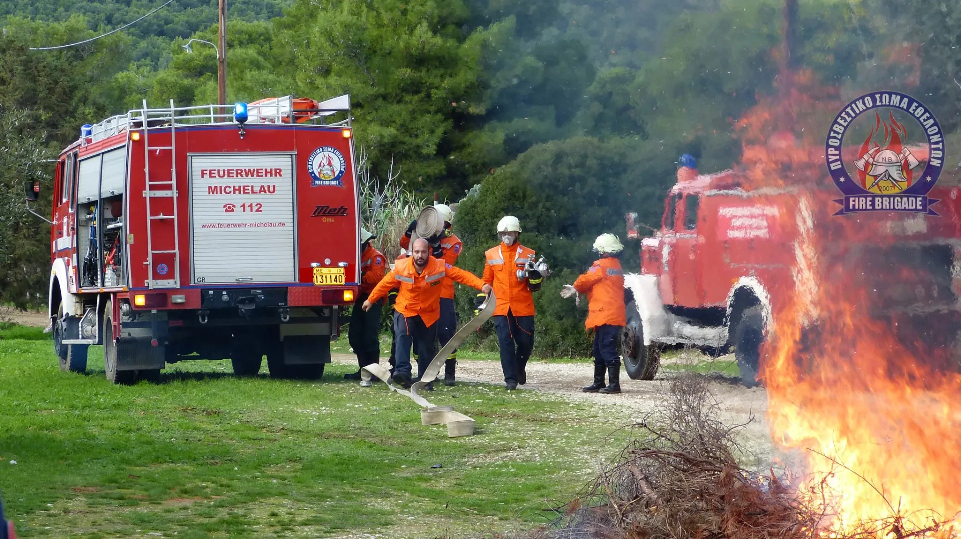 Εθελοντές με στολές κατάσβεσης αντιμετωπίζουν φωτιά σε ξερά χόρτα δίπλα σε πυροσβεστικά οχήματα